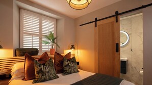 Ein Schlafzimmer mit einem Bett, Kissen, einem Nachttisch, einer Lampe, einer Pflanze, einer Schiebetür und einem Spiegel.