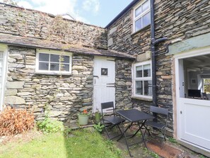 A stone house with a white door, a small patio with a table and chairs, and a garden area with plants and a watering can. - Fell View (Ambleside)