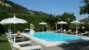 Una piscina con lettini e ombrelloni, immersa nel verde con montagne sullo sfondo.