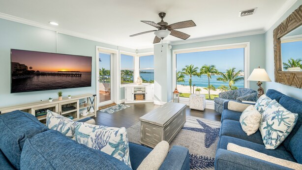 Un soggiorno con una grande TV, divani blu, una ventola a soffitto e vista sulla spiaggia.