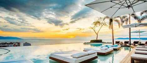 Una piscina infinita con sillones de descanso y sombrillas, con vistas a un atardecer sobre el océano.