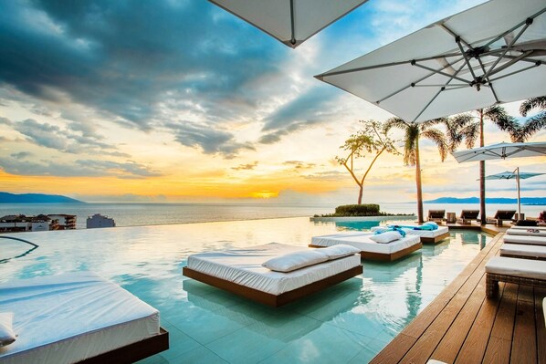 An infinity pool with lounge chairs and umbrellas, overlooking a sunset over the ocean.