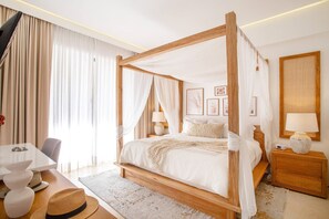 En seng med himmelseng i træ, hvide sengetøj og et natbord med en lampe.