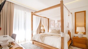 A four-poster bed with white linens, a wooden nightstand with a lamp, a table with hats, and a view of the room's decor.