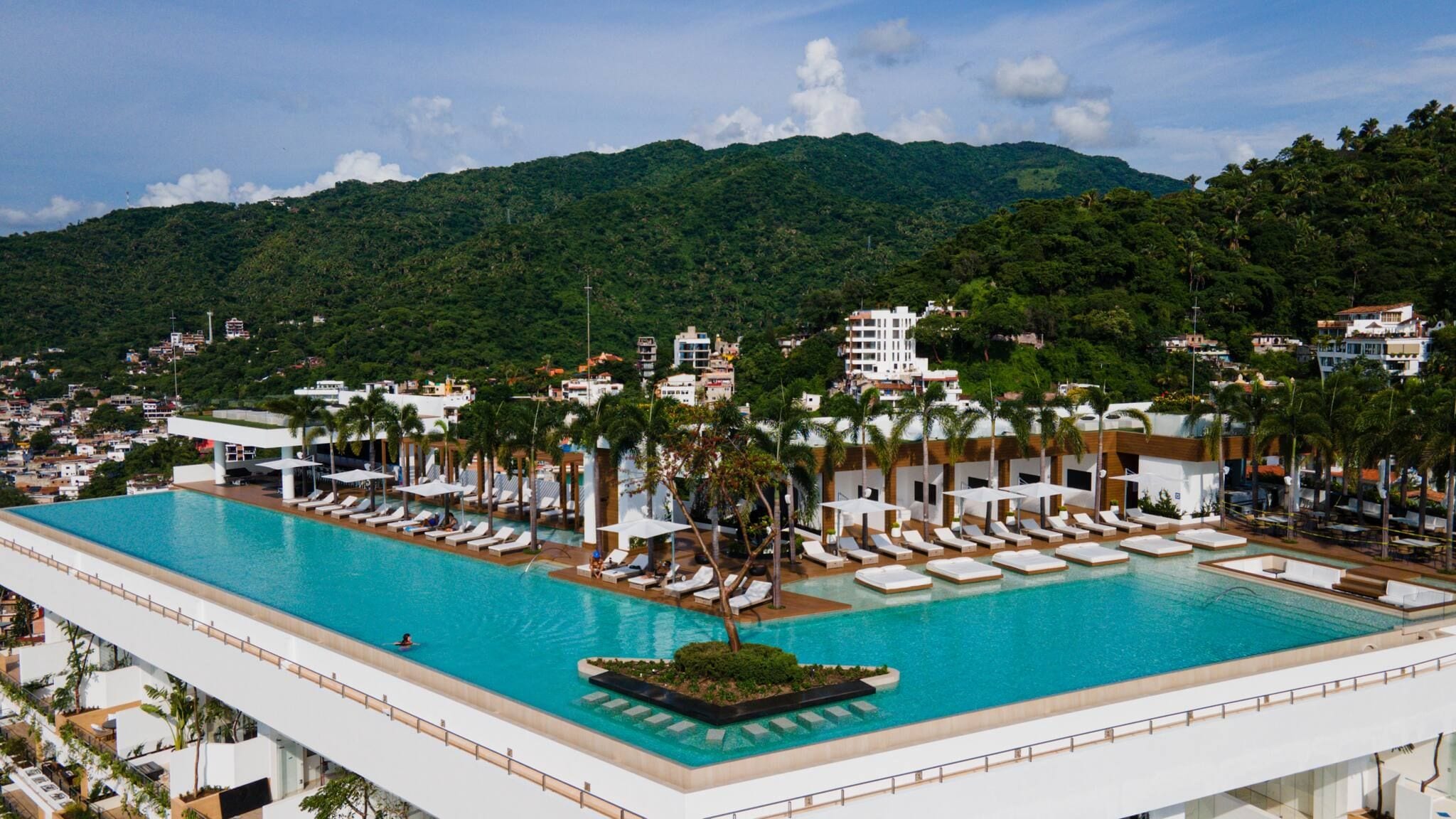 A large outdoor swimming pool with multiple lounge areas and palm trees, surrounded by buildings and a mountainous backdrop.