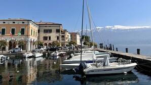 A marina with boats docked, a waterfront building, and a mountain range in the background. - Villa del Lis (Gargnano)