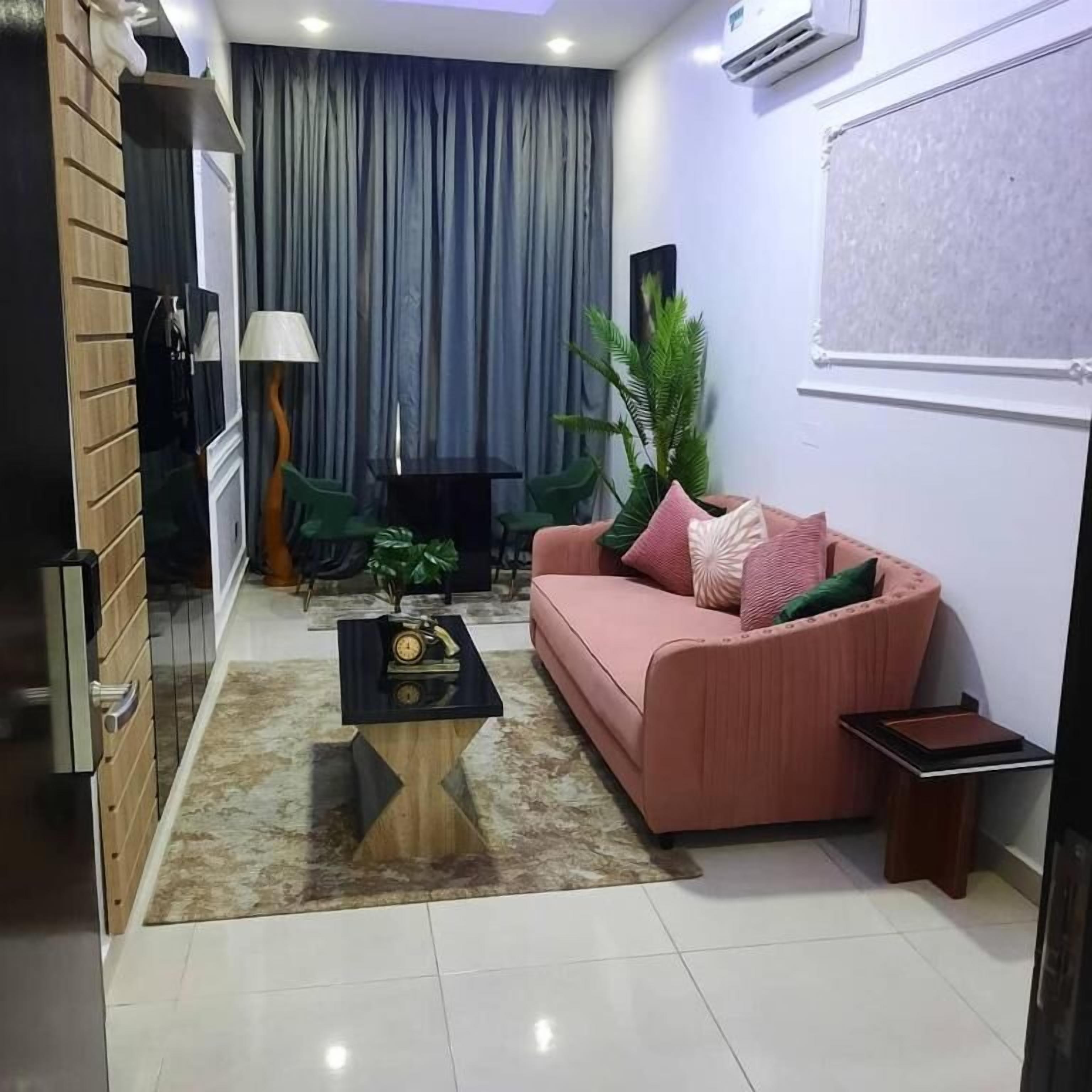 Ein modernes Wohnzimmer mit Sofa, Couchtisch und einem kleinen Beistelltisch. Eine Lampe, eine Uhr und eine Pflanze sind ebenfalls vorhanden. Das Zimmer verfügt über ein großes Fenster mit Vorhängen und eine Klimaanlage an der Wand.
