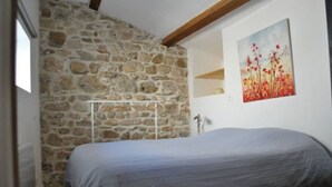 A bedroom with a stone wall, a bed, a painting, and a small shelf. - Les Deux Tours (Aiguèze)