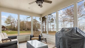 Une véranda avec de grandes fenêtres, un ventilateur de plafond, un barbecue couvert et un coin salon avec une table basse et des chaises.