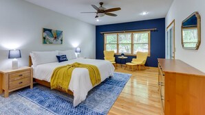 A bedroom with a bed, bedside tables, a ceiling fan, a window with blinds, and a view of trees outside.