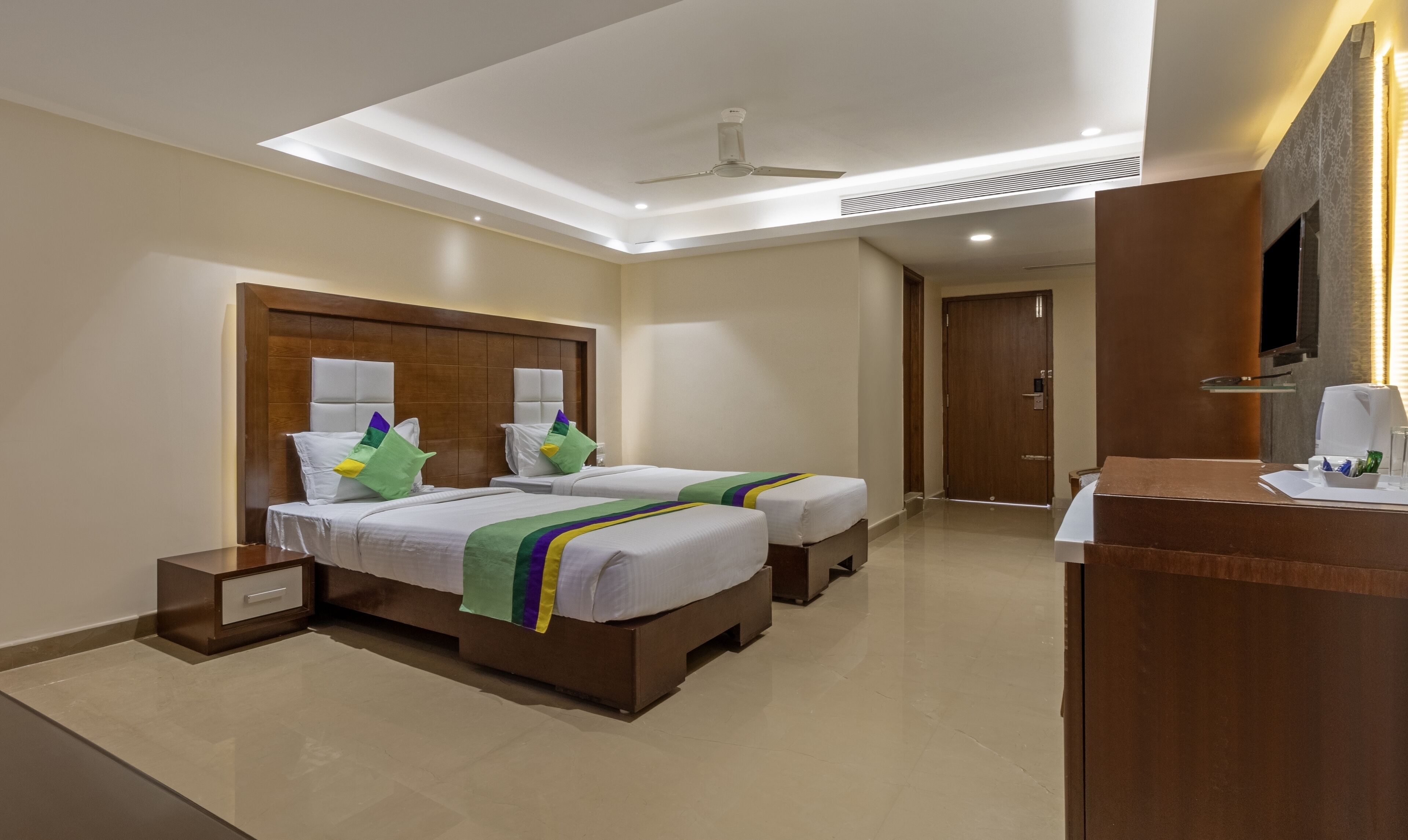 A hotel room with two beds, a wooden headboard, a desk with a chair, a flat-screen TV, and a ceiling fan.