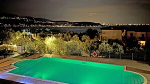 Ein Swimmingpool mit grünlichem Wasser, umgeben von Bäumen und Blick auf eine nächtliche Stadtlandschaft.