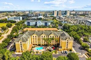 A large hotel with a swimming pool, surrounded by greenery and a cityscape in the background.