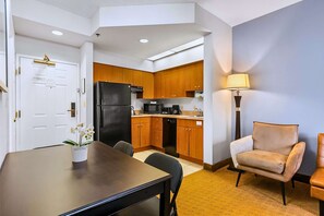 A compact kitchen with a refrigerator, microwave, and coffee maker, a dining table with chairs, and a lamp.