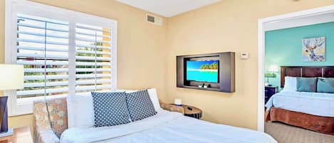 Ein Hotelzimmer mit einem Bett, einem an der Wand montierten Fernseher, einem Fenster mit Jalousien und einer Tür zu einem weiteren Raum.