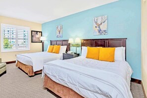 A hotel room with two beds, a window with blinds, a nightstand, and artwork on the walls.
