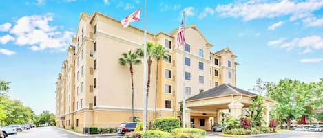 A multi-story hotel with a beige facade, palm trees, and two flags.