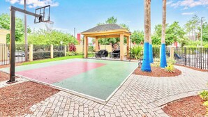Un terrain de basket avec un kiosque, des palmiers et une zone délimitée par une clôture.