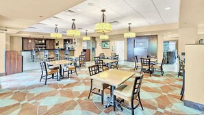 Un grand espace dédié à la salle à manger, avec plusieurs tables et chaises, une cuisine à l’arrière-plan et un tapis à motifs.