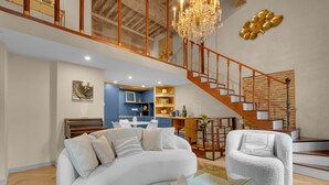 Un vaste espace de vie comprenant un canapé blanc incurvé, une table basse en verre et un fauteuil blanc. Un escalier en bois, doté d’une rampe, mène à un niveau supérieur.