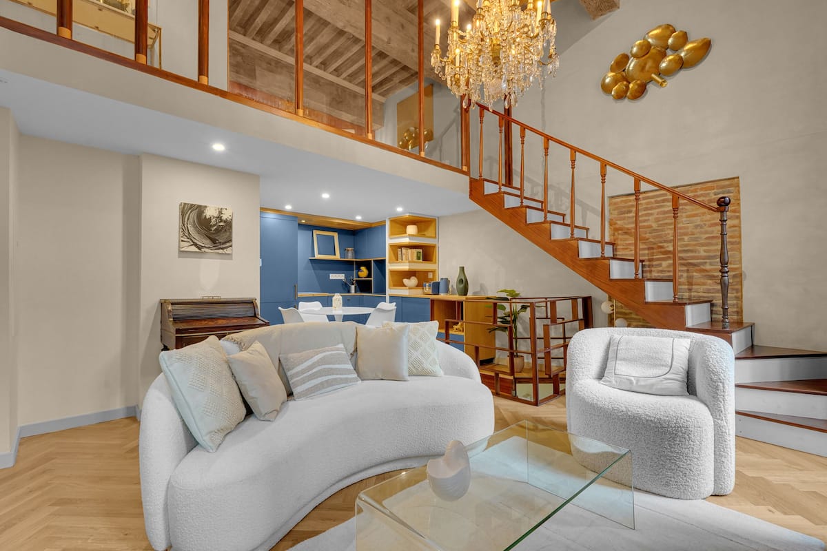 A spacious living area with a curved white sofa, a glass coffee table, and a white armchair. A wooden staircase with a railing leads to an upper level.