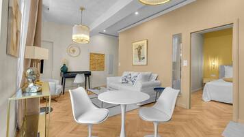 A modern living room with a white sofa, a round coffee table, and two white chairs. There is a desk with a lamp and a decorative bust on the left. A painting and a framed picture adorn the walls.