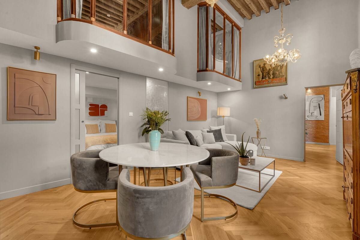 A modern living room with a round white table, grey armchairs, a sofa, and a chandelier.