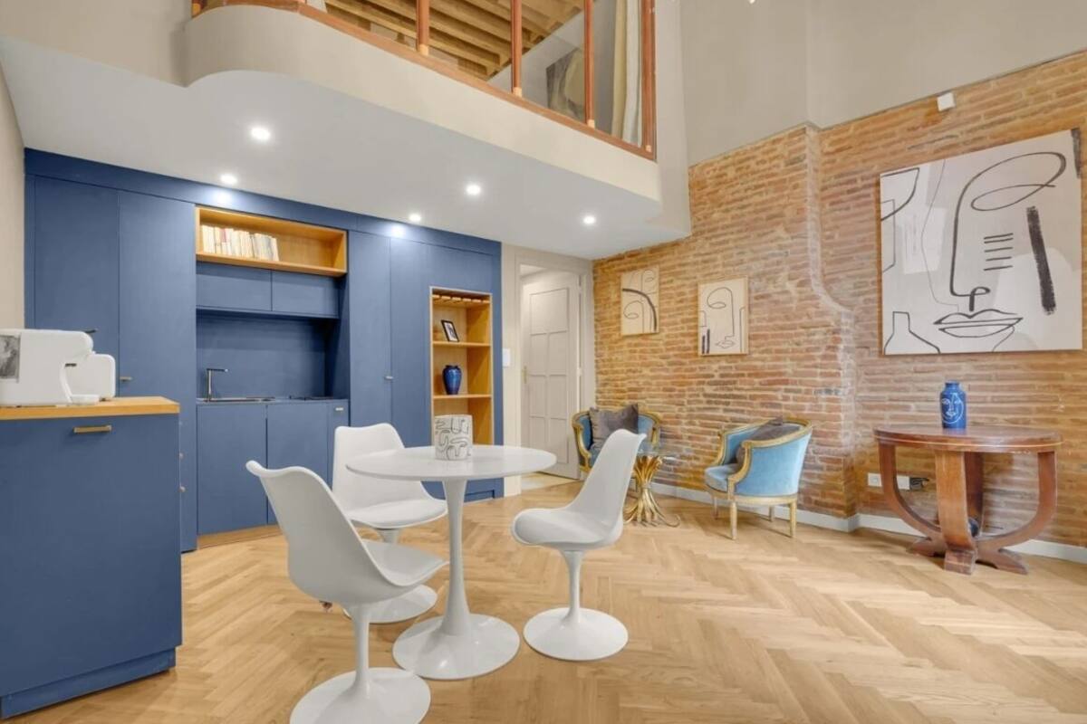 A modern kitchen with blue cabinets, a dining area with white chairs, and a brick wall with artwork.