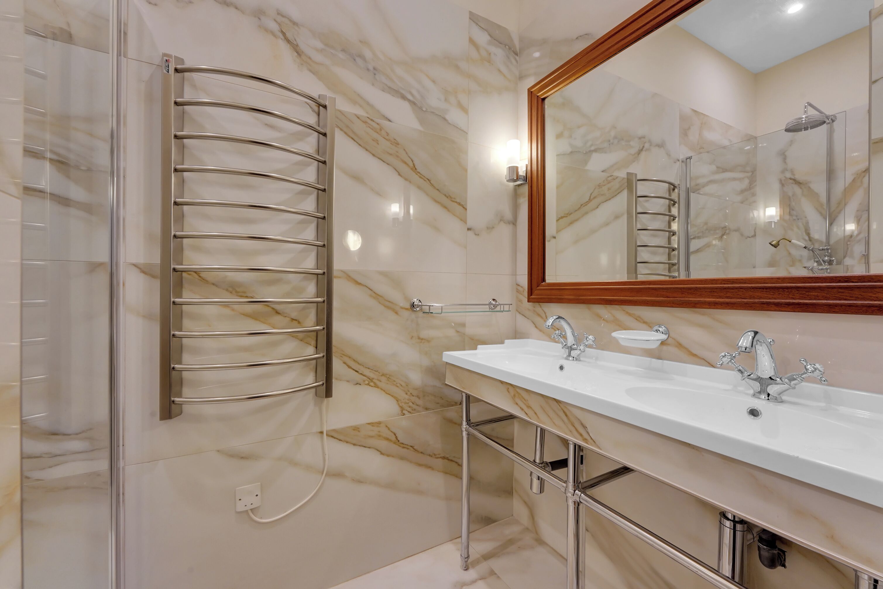 A modern bathroom with a glass shower enclosure, a marble-tiled wall, a double sink with chrome fixtures, and a large mirror.