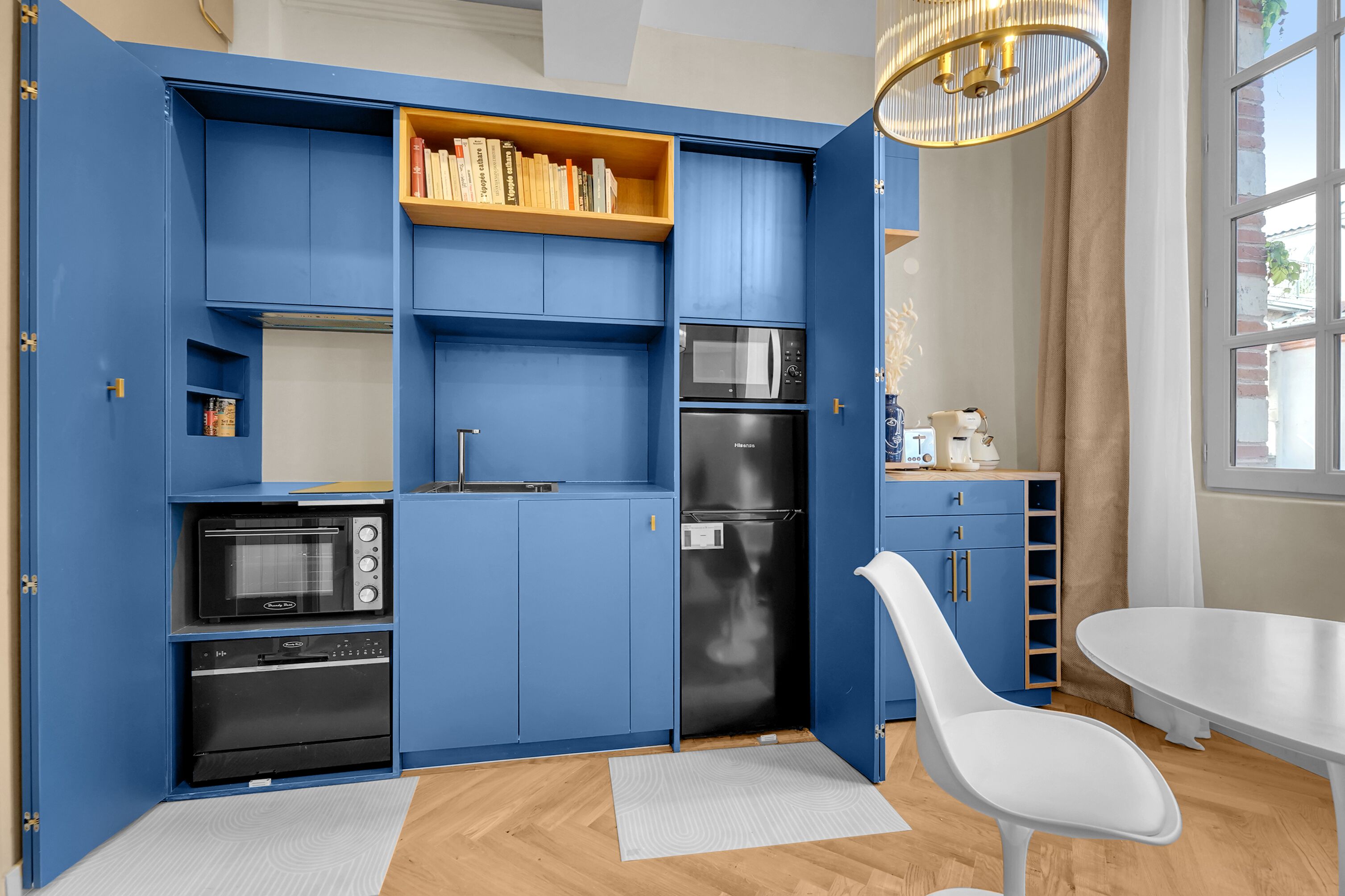 Eine moderne Küche mit blauen Schränken, einem eingebauten Backofen, einer Mikrowelle und einem Kühlschrank. Ein weißer Stuhl und ein kleiner Esstisch sind ebenfalls vorhanden.