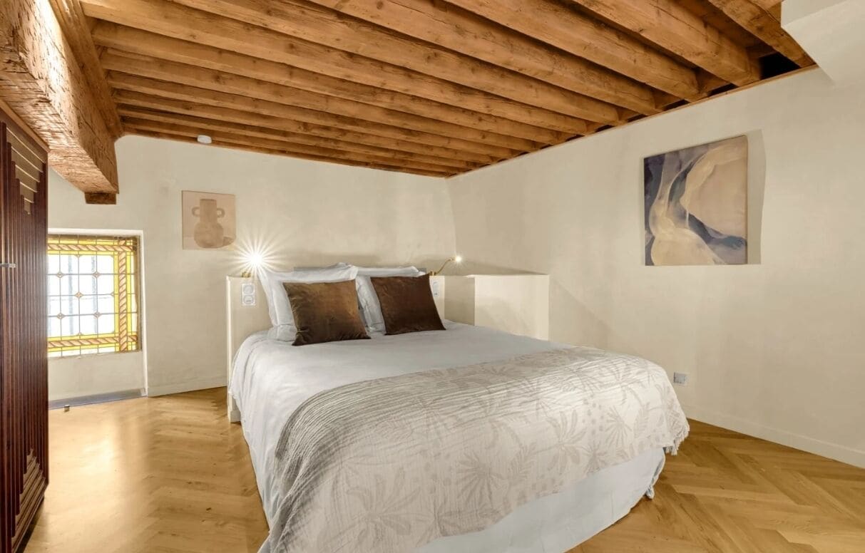 A bedroom with a wooden ceiling, a bed with a patterned bedspread, a window with bars, and a painting on the wall.