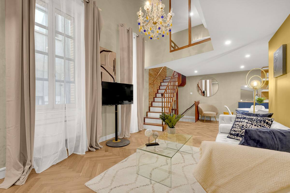 A modern living room with a large window, a flat-screen TV, a glass coffee table, a staircase, and a chandelier.