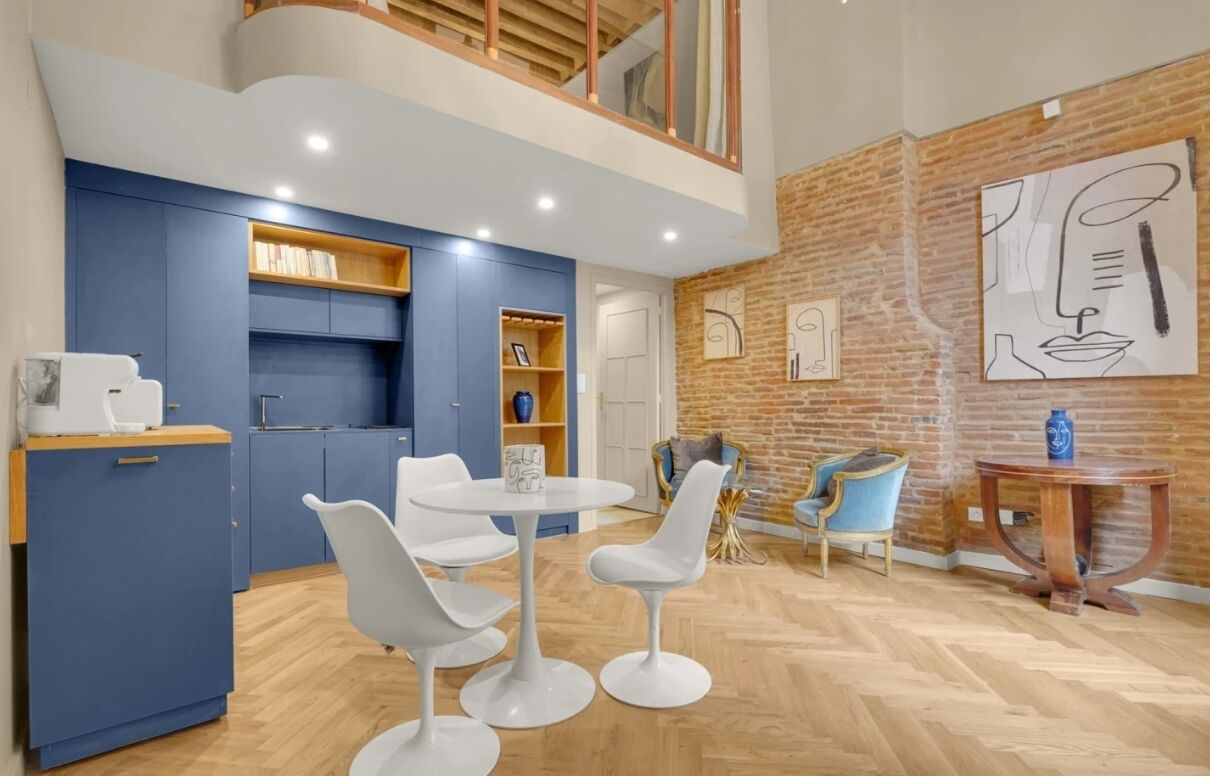 A modern kitchen with blue cabinets, a dining area with white chairs, and a brick wall with abstract art.
