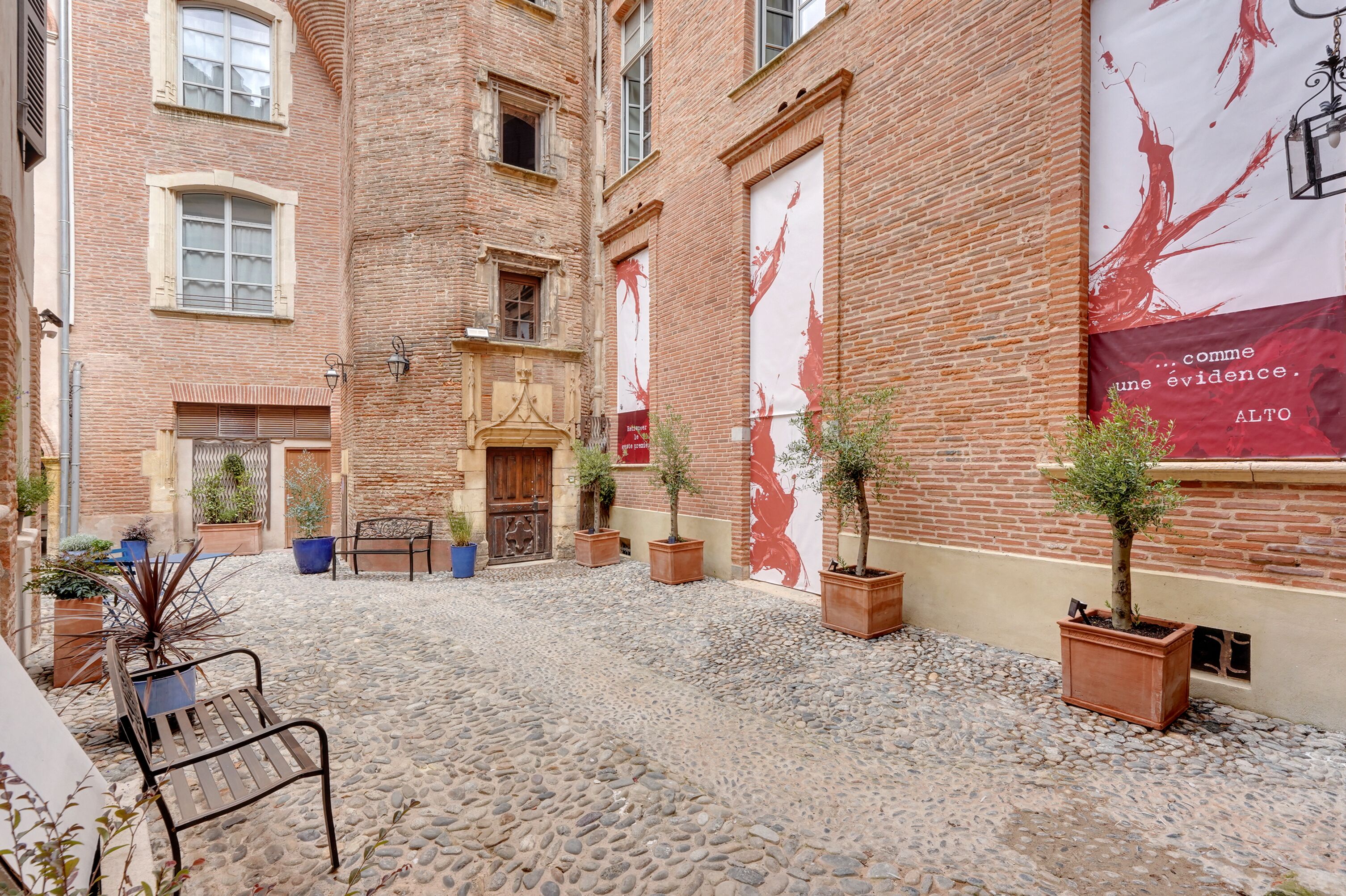 A courtyard with a cobblestone floor, potted plants, a bench, and a building with a large red banner.