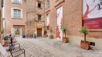 A courtyard with a cobblestone floor, potted plants, a bench, and a building with a large red banner.