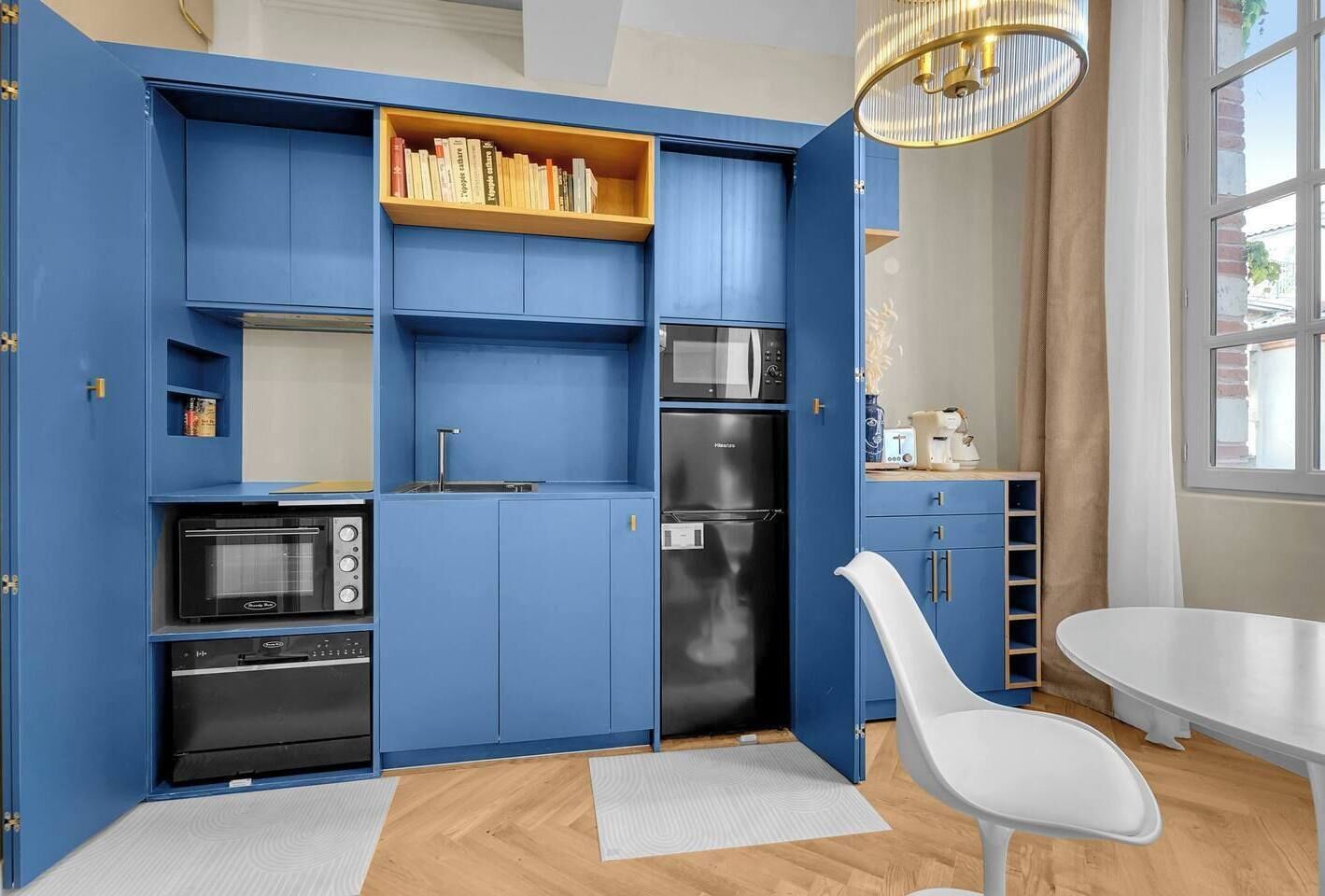 Una cocina compacta con armarios azules, un refrigerador negro, un microondas y un horno empotrado. Hay una silla blanca y una pequeña mesa de comedor.