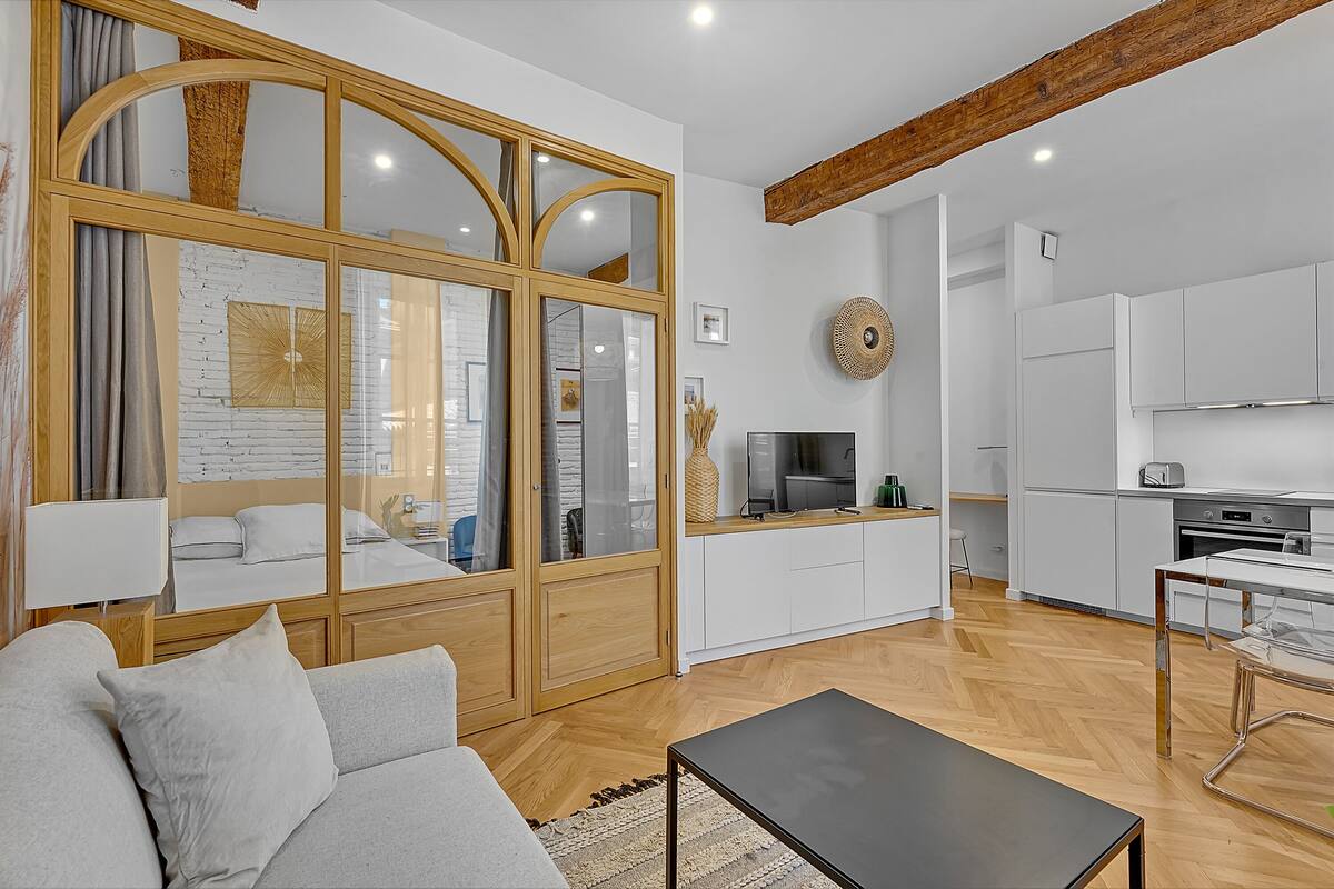 A modern living room with a sofa, a coffee table, and a TV. There is a kitchen area with cabinets and a dining table.