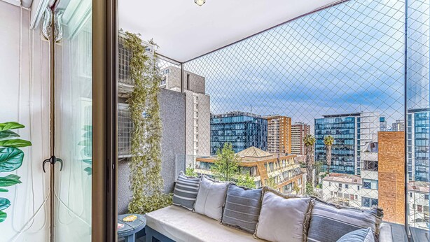 A balcony with a cushioned seating area, a small table with a plate, and a view of the cityscape through a mesh window. - Stylish Art 2B w/ Balcony, Parking & Wifi (Santiago)
