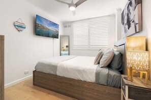A bedroom with a bed, a nightstand, a wall-mounted TV, and a window with blinds. - Stylish Manly Apartment with Harbour Views (Manly)