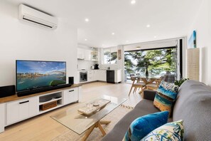 A modern living room with a flat-screen TV, a glass coffee table, and a sofa with colorful cushions. - Stylish Manly Apartment with Harbour Views (Manly)