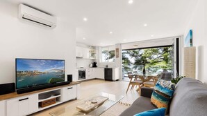 A modern living room with a flat-screen TV, a glass coffee table, and a grey sofa with colorful cushions. A kitchen with white cabinets and a dining area with a glass table and chairs is visible through an open door. - Stylish Manly Apartment with Harbour Views (Manly)