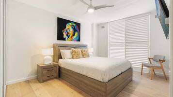 A bedroom with a bed, nightstand, lamp, chair, ceiling fan, and a colorful painting on the wall.