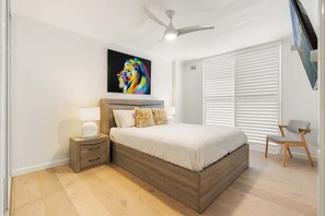 A bedroom with a bed, nightstand, lamp, chair, ceiling fan, and a colorful painting on the wall.