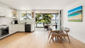 A modern kitchen with white cabinets, a dining area with a glass table, and a large window offering a view of a tree and a beach. - Stylish Manly Apartment with Harbour Views (Manly)