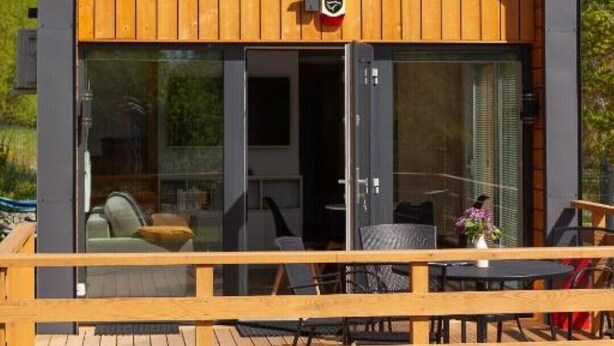 Une cabane moderne dotée d’une terrasse en bois, de grandes fenêtres et d’un logo sur la porte.