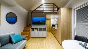 Un espace de vie compact comprenant un canapé, un téléviseur fixé au mur, un escalier en bois et une petite cuisine.