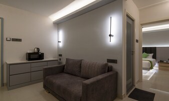 A modern hotel room with a grey sofa, a small kitchenette, and a bed with a headboard.