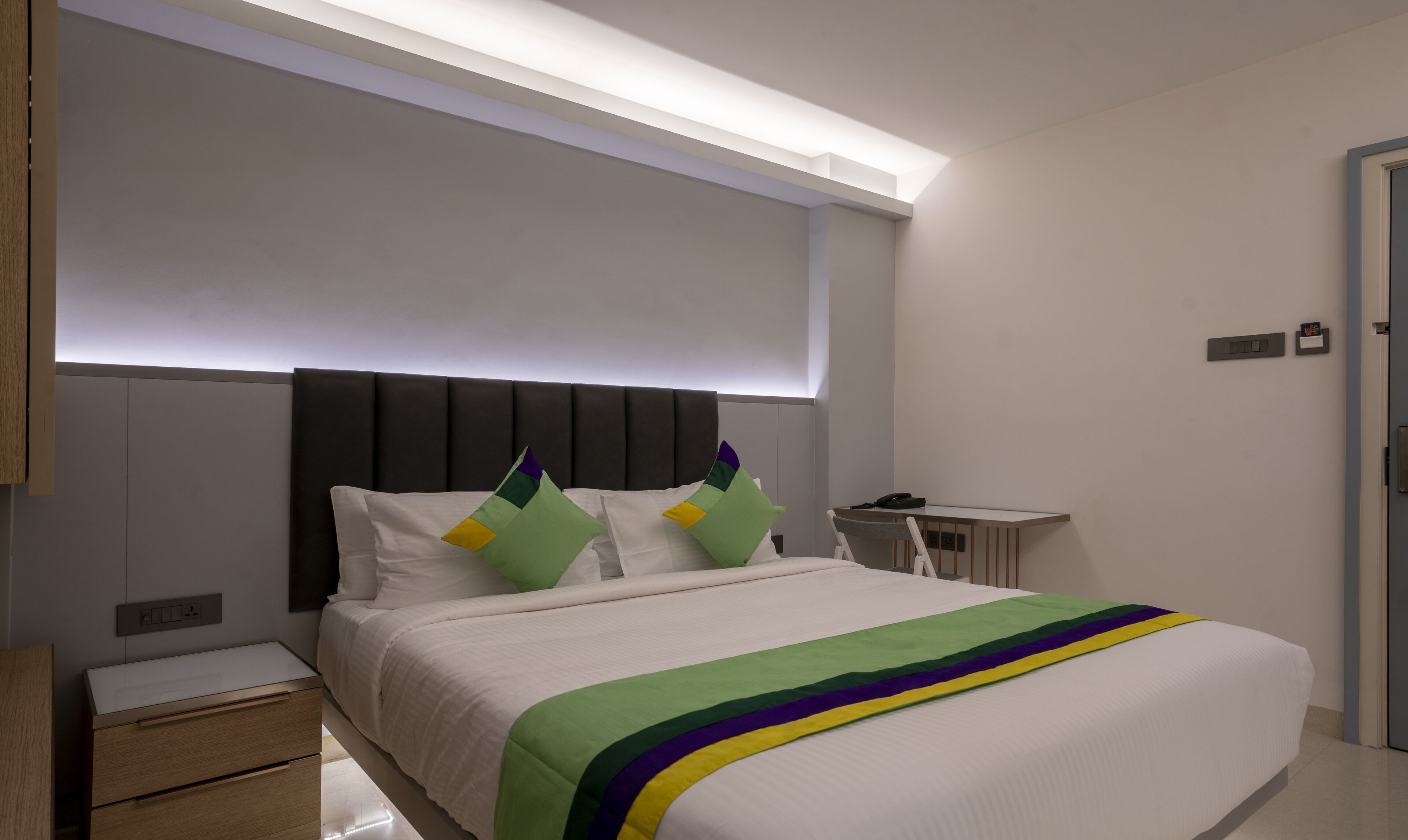 Een moderne hotelkamer met een groot bed, nachtkastje, bureau en een hoofdbord met ingebouwde verlichting.