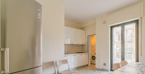 Een kleine keuken met witte kastjes, een eettafel met stoelen en een betegelde vloer.
