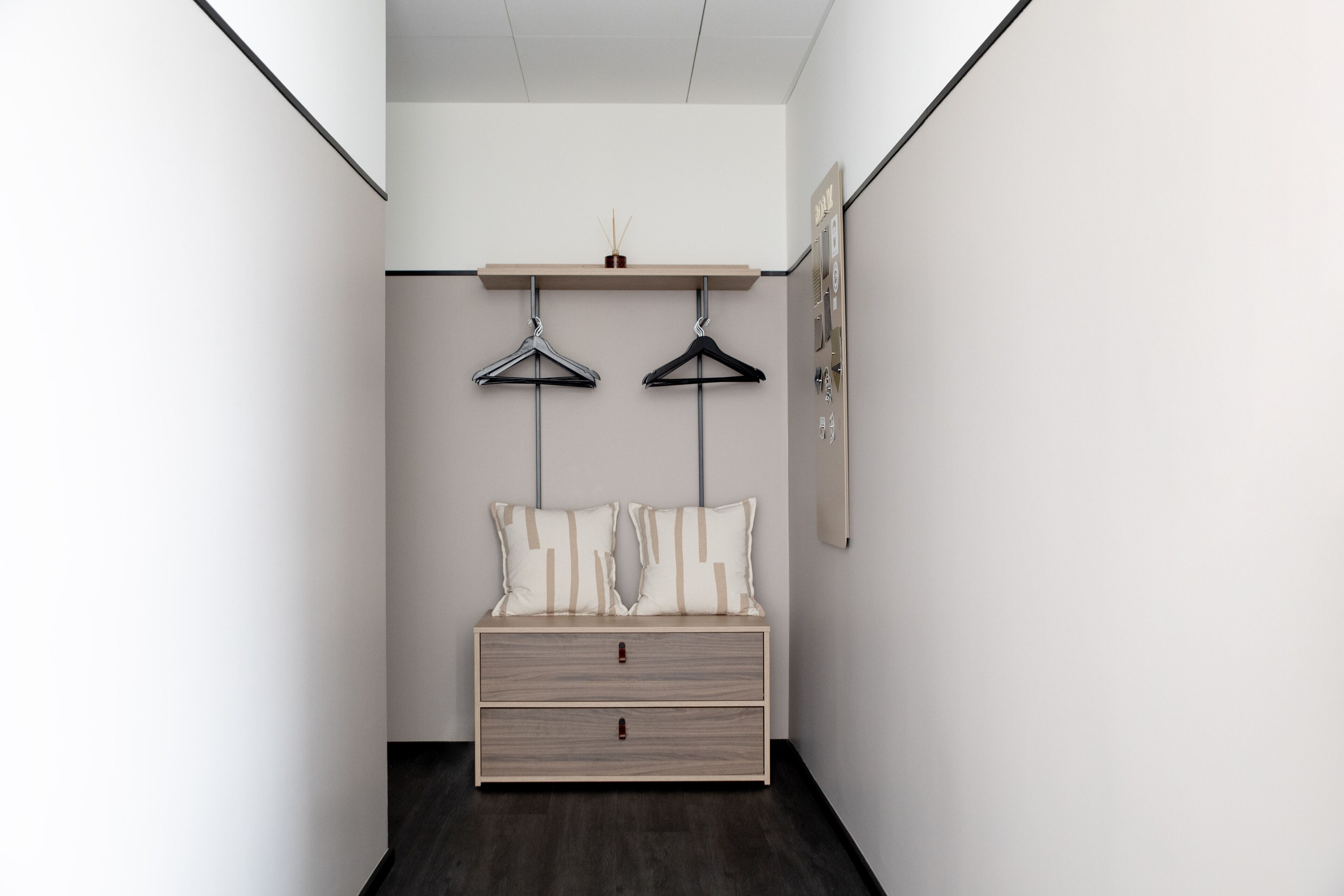 A narrow hallway with a bed at the end, a hanging shelf above, and a framed picture on the wall.